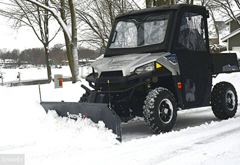 UTV (Utility Terrain Vehicle) with a cab equipped with a SnowEx snow plow actively clearing snow in winter on a snow-covered residential street. 