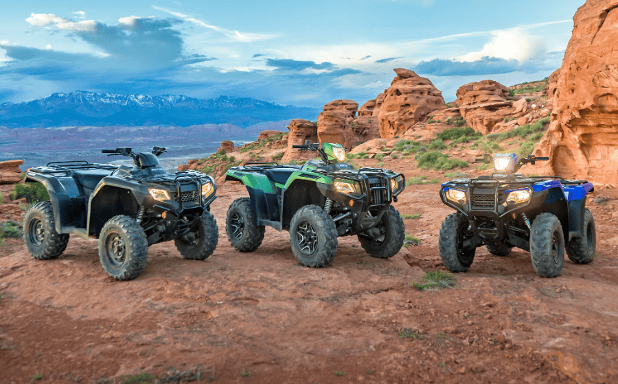 Three ATVs Parked Side By Side on a Dusty Trail at Dusk, With Rocky Desert Formations in the Background and Distant Mountains Under a Soft, Cloudy Sky