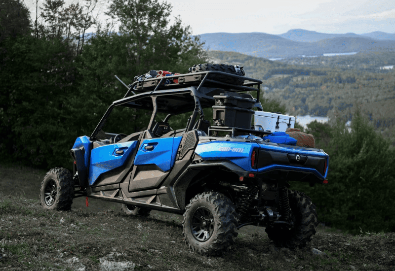 Side Profile of a Blue UTV Parked On a Grassy Hillside Set Against a Backdrop of Distant Mountains, Bodies of Water, and Forests