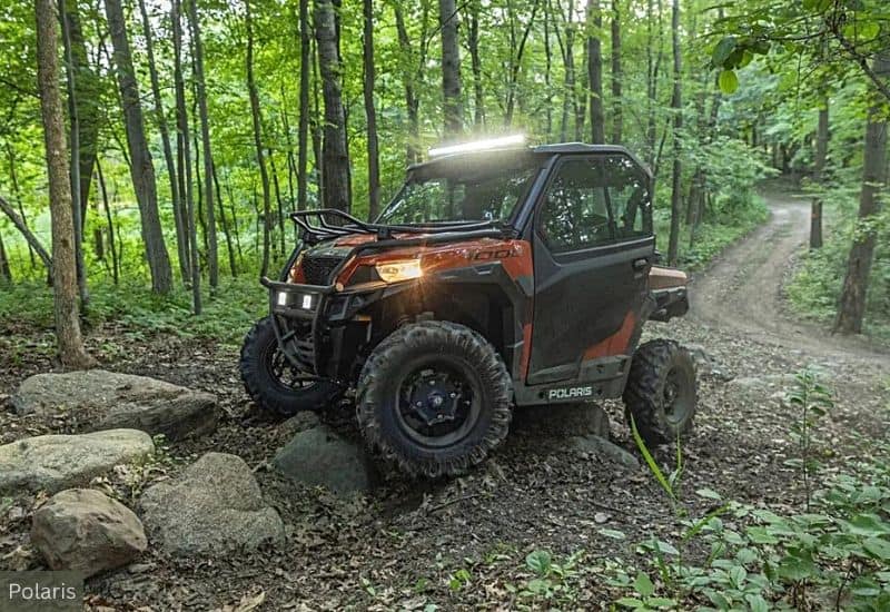 Black and Orange Polaris UTV Navigating Through a Forest Trail With its Lights On Climbing Over a Large Rock