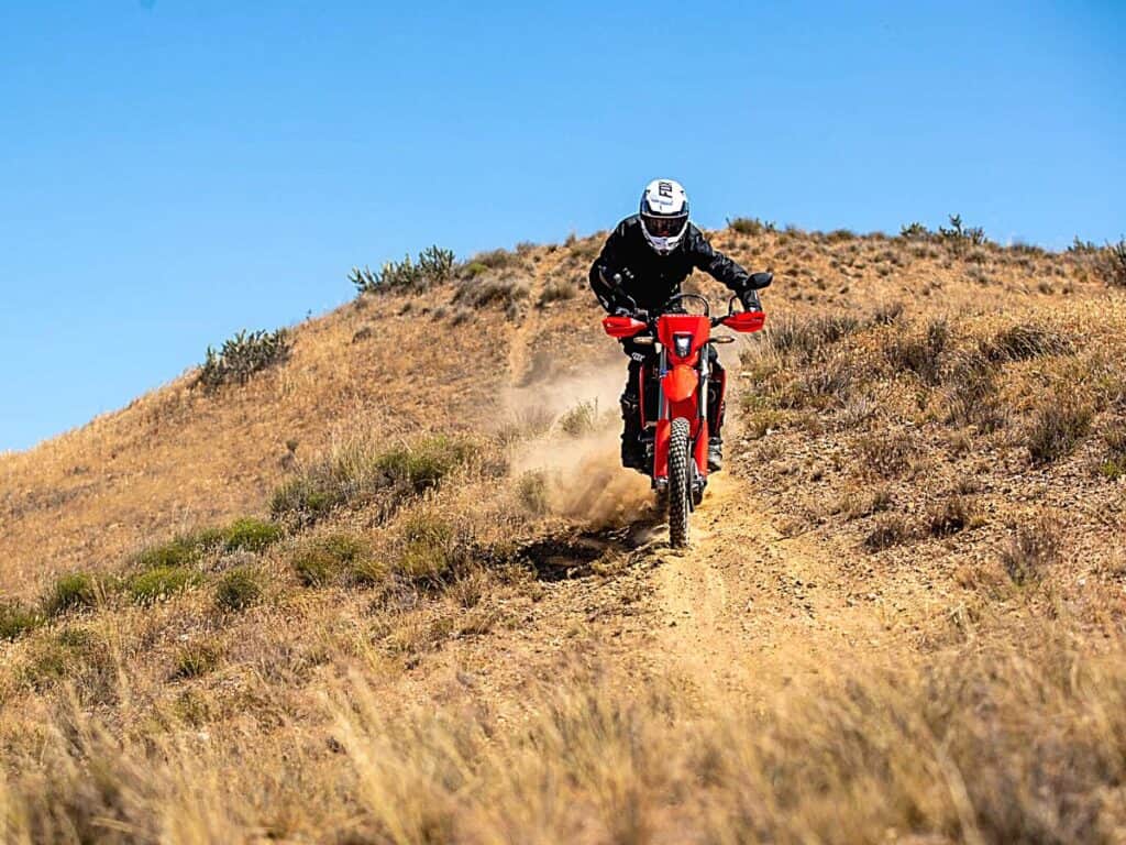 Rider Clad in Black Riding Gear is Maneuvering a Honda CRF450RL Dual-Sport Motorcycle on a Dusty Trail Over a Sunbaked Hillside.