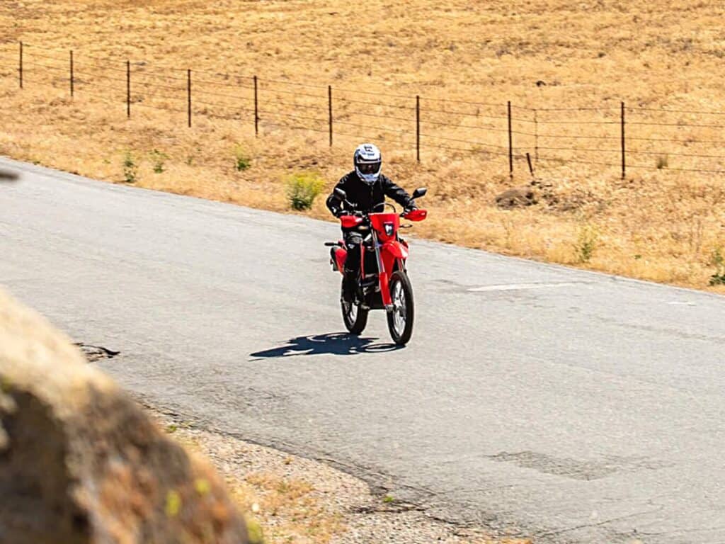 Rider Wearing a Black Jacket and a White Helmet is Piloting a Red Honda CRF450RL Dual-sport Bike on an Asphalt Road