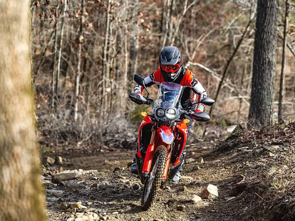 Motorcyclist in a Red, Black, and White Riding Suit Riding a Honda CRF300L Rally Motorcycle on a Rocky Forest Trail