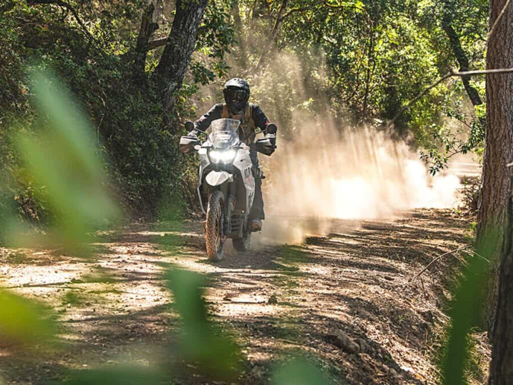 Rider Clad in Black Protective Gear and a Helmet is Riding a Kawasaki KLR 650 Adventure Motorcycle Along a Sun-dappled Dirt Road Surrounded by Dense Greenery