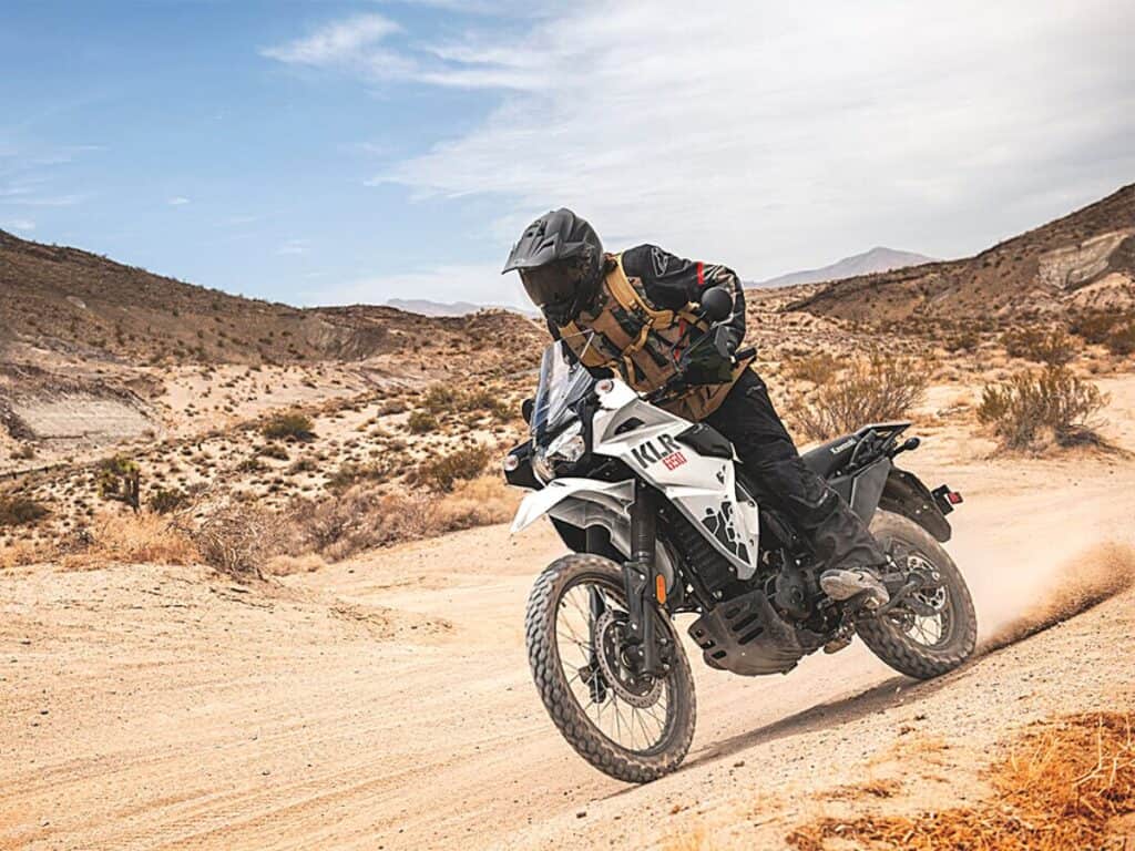Rider Dressed in Black Protective Gear With a Matching Helmet is riding a White Kawasaki KLR 650 Dual Sport Bike on a Dusty Trail in a Desert Landscape