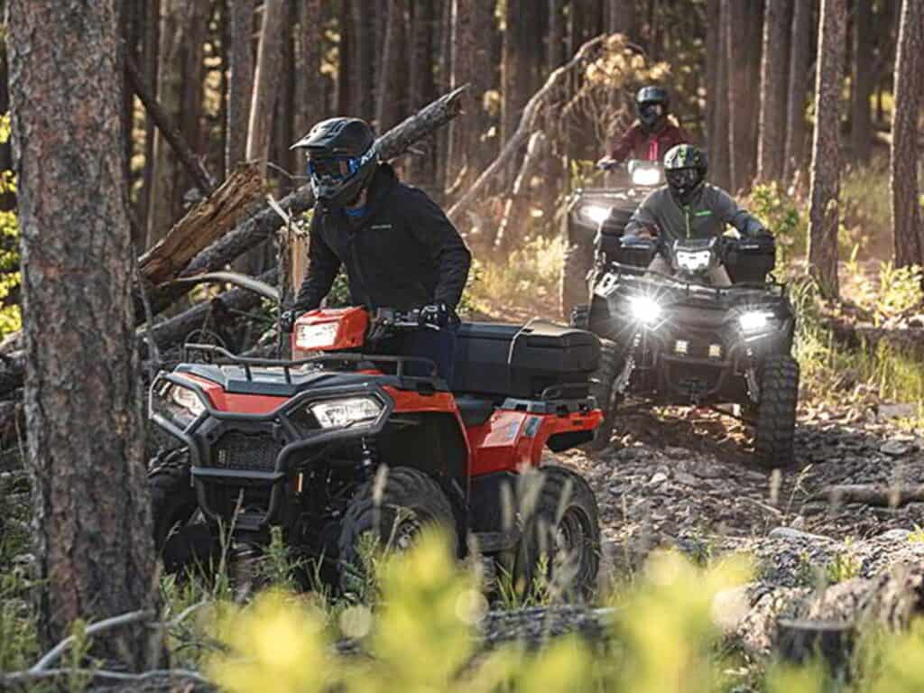 Three Riders Navigating Through a Dense Forest Trail on Polaris Sportsman 570 ATVs With Their Headlights on