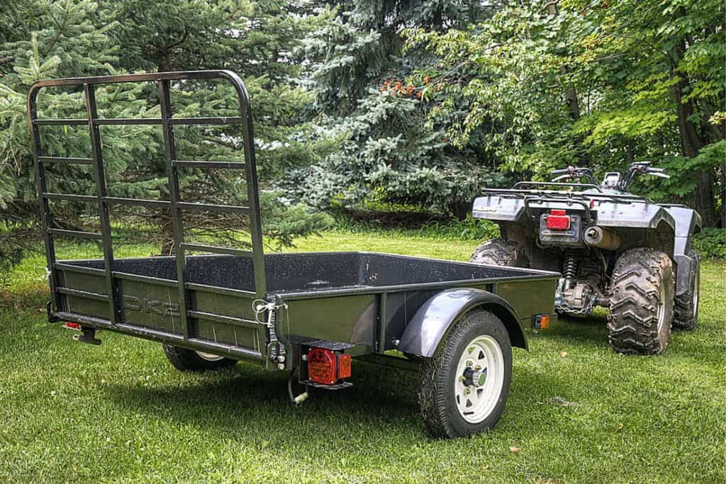 Single-axle Utility Trailer with a Tall Mesh Tailgate Parked on Grass, with an ATV in the Background Positioned as if Ready to be Hitched to the Trailer