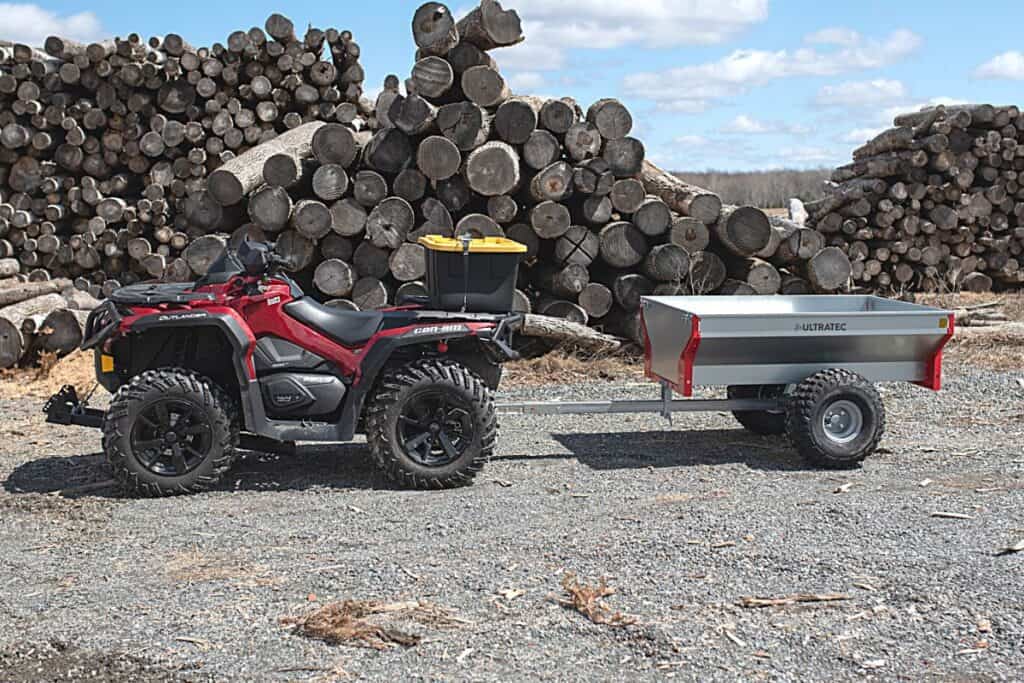 Red ATV Connected to a Metal Single-axle Utility Trailer Parked on a Gravel Lot, with a Large Stack of Cut Logs in the Background