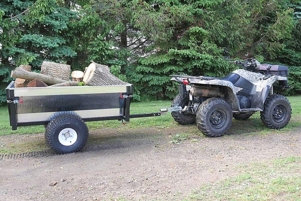 Loaded Single-axle Utility Trailer with Logs, Hitched to a Muddy ATV, Parked in a Grassy Area with Trees in the Background