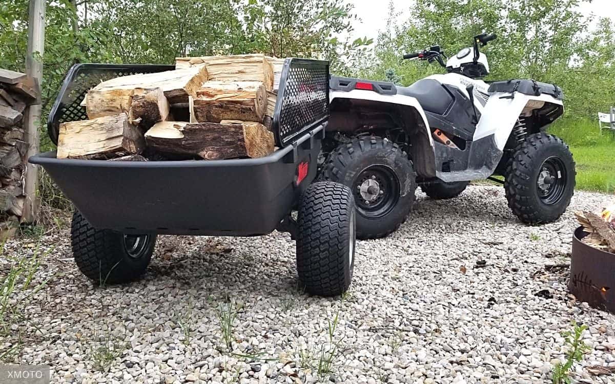 Black, Single-axle Dump Trailer Loaded with cut Firewood Attached to a White ATV on a Gravel Surface, Surrounded by Greenery