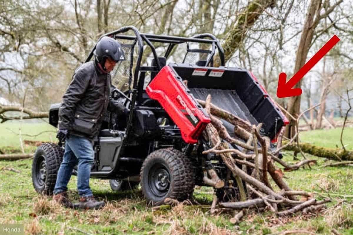 A person is unloading branches from the tiltable cargo bed of a Honda Pioneer UTV in a wooded area.