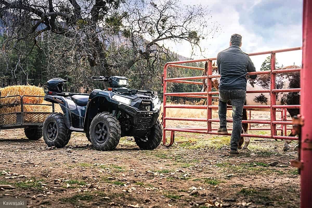 A Kawasaki Brute Force 750 ATV is parked next to a hay-laden trailer within a rustic farm setting. A man stands facing away from the camera, leaning on a red metal gate while observing a horse beyond the fence, indicating the ATV's utility role in farm life. The environment is natural and serene, with bare trees and a calm sky in the background, suggesting a cool or overcast day.