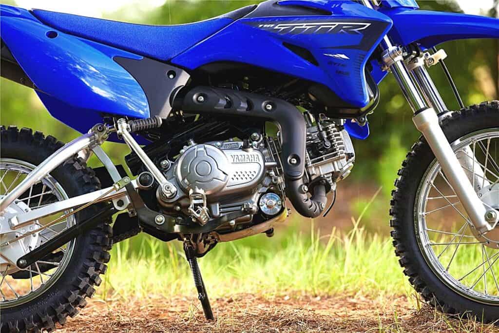 A close-up view of a Yamaha TT-R110 dirt bike, focusing on its midsection. The bike's vibrant blue bodywork is adorned with white 'TT-R' graphics, and the Yamaha logo is visible on the engine case. The engine itself appears compact and is air-cooled, with a black exhaust pipe that curves around the side. The bike stands on a kickstand, highlighting the knobby off-road tires and sturdy suspension components, which indicate its capability for trail riding. The background is softly blurred, emphasizing the dirt bike's details and craftsmanship.