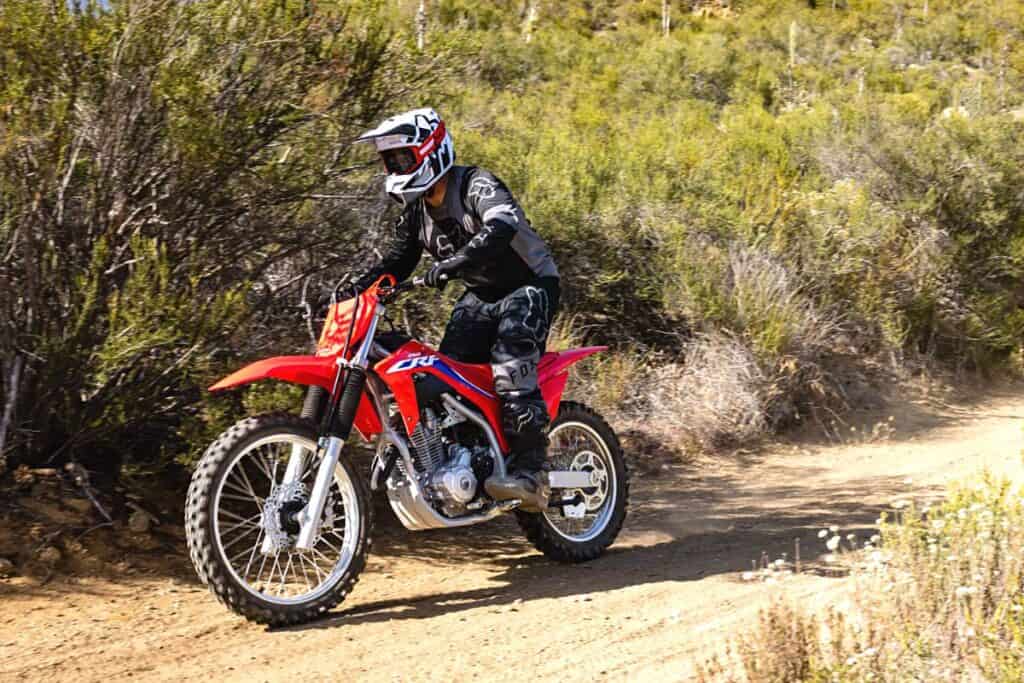Rider on an air-cooled Honda CRF250F, maneuvering on a dirt trail. The rider is wearing full gear, including a black and white helmet, gloves, and protective clothing. The bike is in motion, over uneven terrain. The setting is a natural, shrub-lined trail that suggests the bike is being used for trail riding.