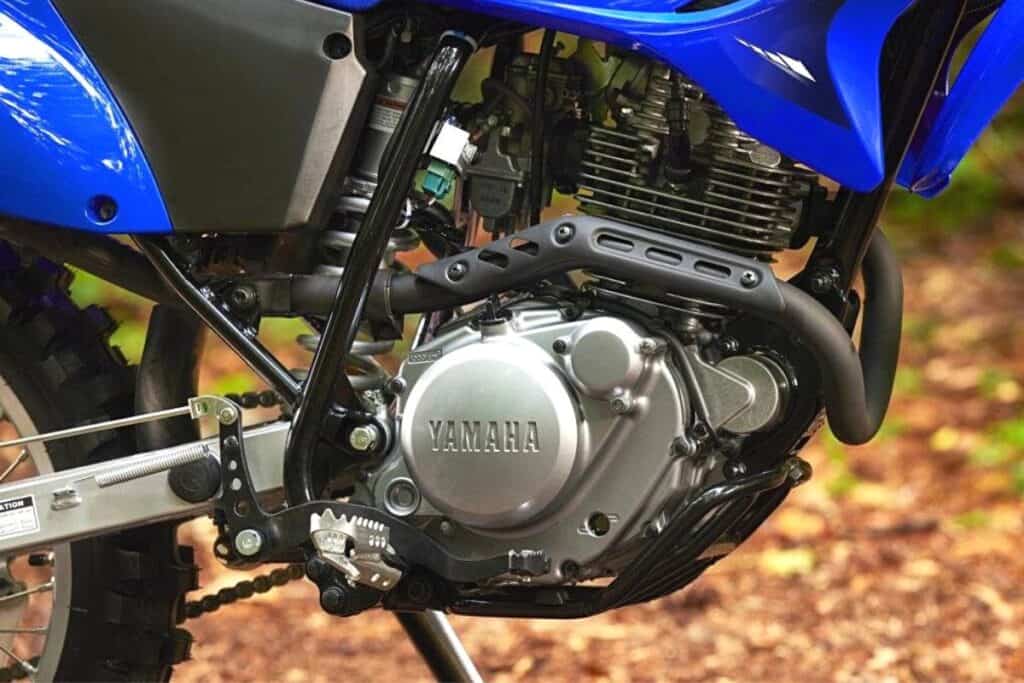 A close-up of the engine and lower chassis of a Yamaha TT-R230 dirt bike, highlighting its air-cooled engine design. The bike's blue plastics partially cover the upper area of the engine, which features the Yamaha logo prominently on the engine casing. The details of the engine fins, which aid in dissipating heat, are visible, indicating the air-cooling system.