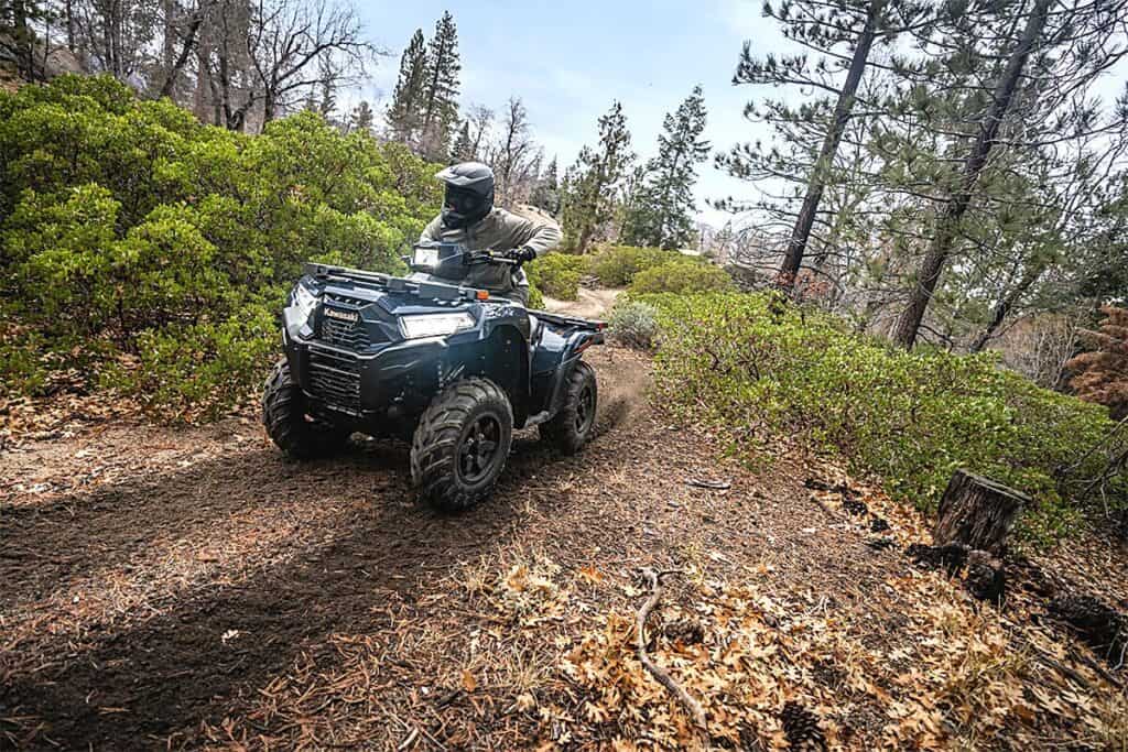 A rider atop a Kawasaki Brute Force 750 is traversing a forest trail, showcasing the vehicle's off-road prowess. The ATV is navigating a path with loose soil and surrounded by shrubs and pine trees, illustrating the rugged terrain it is capable of handling.