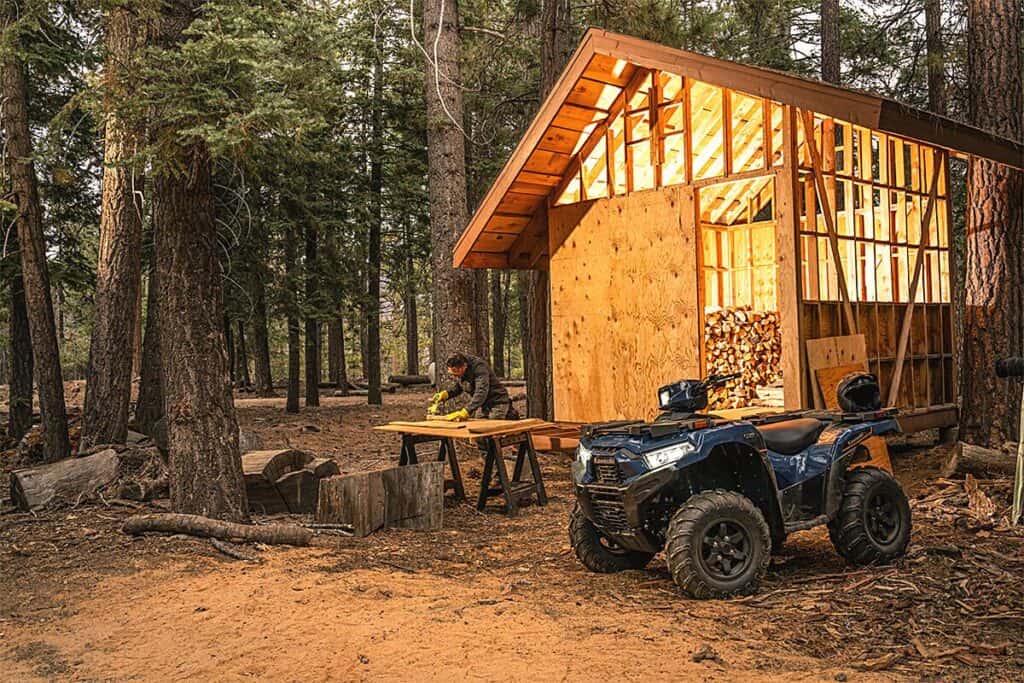 In a serene woodland setting, a Kawasaki Brute Force 750 EPS 4x4i EPS ATV stands to the right of the frame, showcasing its rugged design suited for outdoor tasks. To the left, a person wearing a jacket and gloves is actively working. Behind them is a partially constructed wooden cabin with a neatly stacked woodpile under its sheltering overhang, implying the use of the ATV in aiding construction or wood-gathering efforts. The scene is enveloped by tall, mature trees, suggesting a remote or wilderness area.