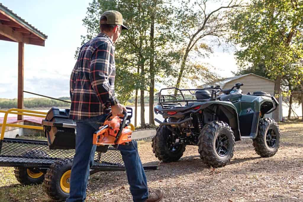 A man in a plaid shirt and baseball cap is walking towards an ATV, carrying an orange chainsaw. In the background, a Can-Am Outlander ATV is parked. The ATV is equipped with large, aggressive tires and appears ready for robust outdoor activity. Behind the ATV, there's a utility trailer, indicating its use for hauling equipment. The setting is outdoors with trees, a large body of water, and a clear sky, which implies a rural or semi-rural location with open spaces suitable for ATV use and outdoor work.