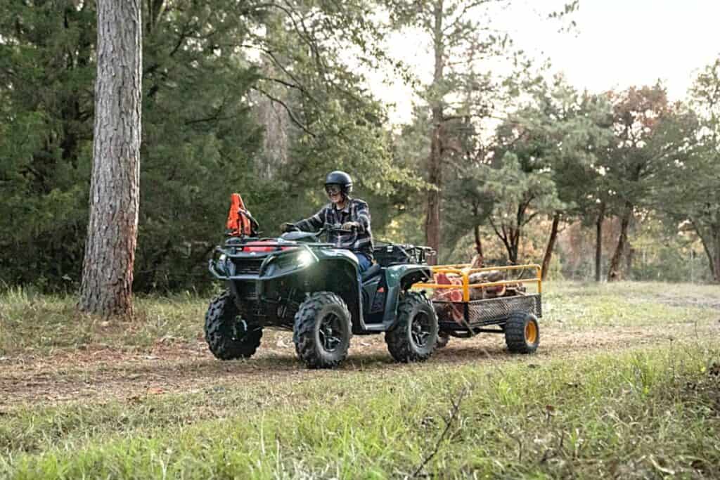 A rider wearing a helmet and plaid shirt is seated on a Can-Am Outlander ATV, towing a small utility trailer loaded with wood.