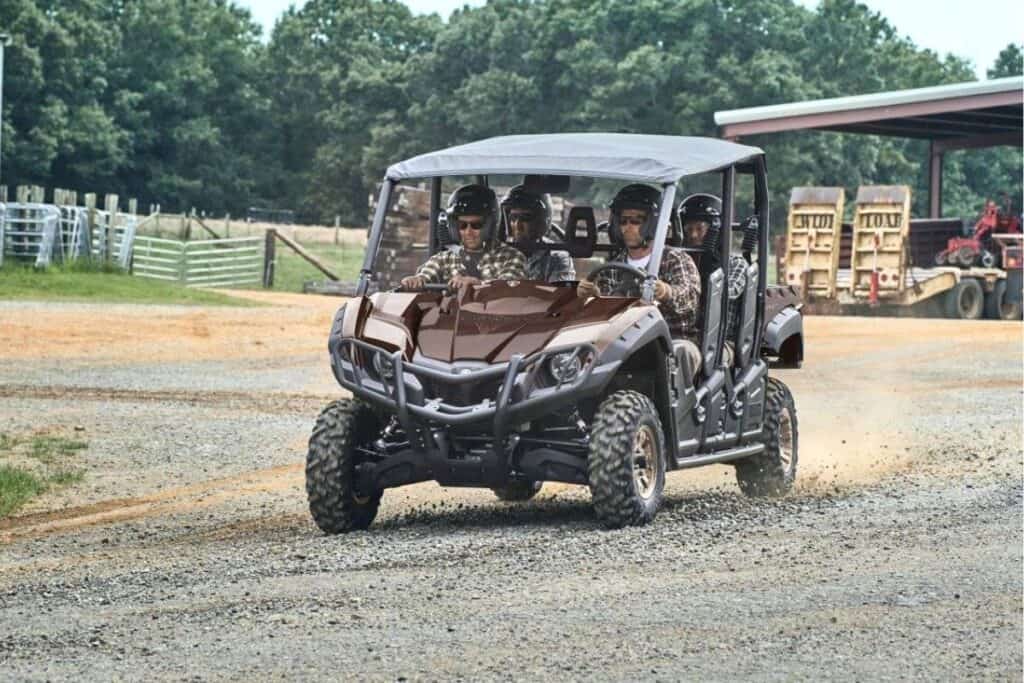 A group of riders in a Yamaha Viking VI EPS UTV are driving down a gravel path on a farm, with fences and farm equipment in the background.