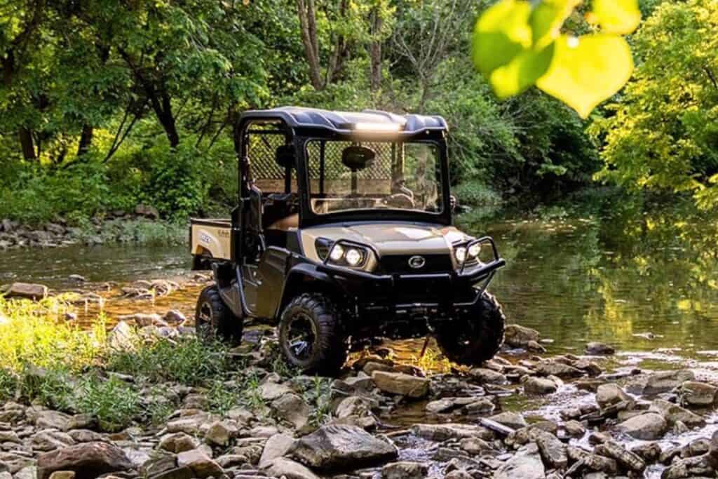 A Kubota Sidekick RTV-XG850 UTV parked by a creek in a forest, reflecting the sunlight on the water's surface.