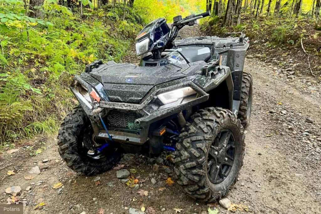 A Polaris Sportsman XP 1000 ATV covered in mud on a forest trail.