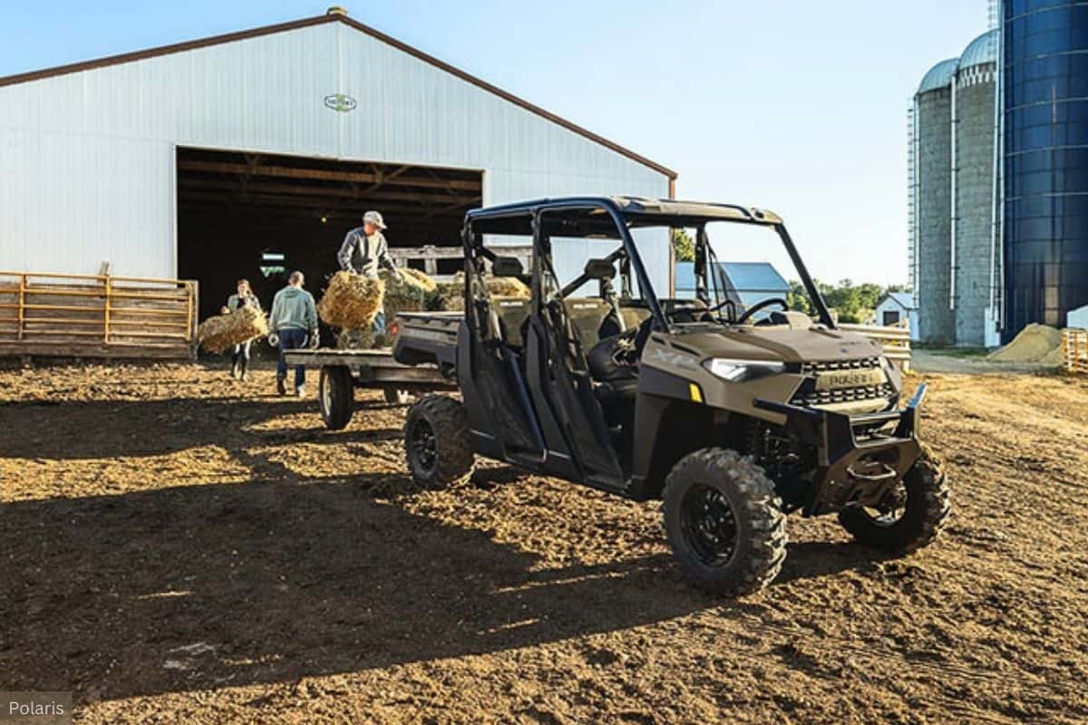 Side-by-side utility terrain vehicle (UTV) made by Polaris parked in front of a barn with a silo in the background. There are three people in the image; one is standing on the trailer being pulled by the UTV, and two are on the ground, all handling hay.