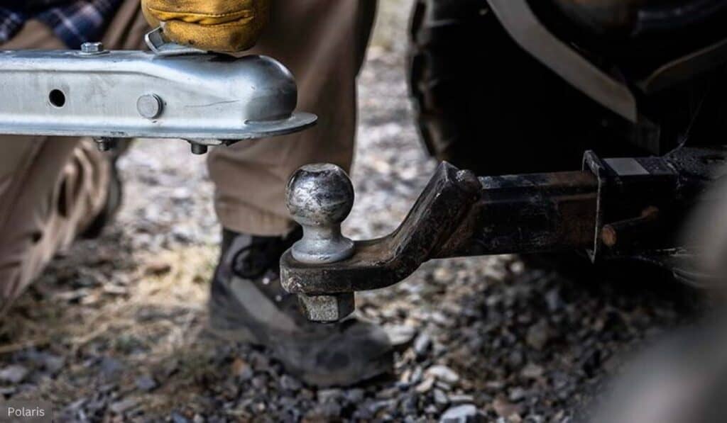 Close-up view of a hitch ball on a UTV. The hand of a person, wearing a leather work glove, is guiding the trailer hitch receiver towards the hitch ball to connect them.