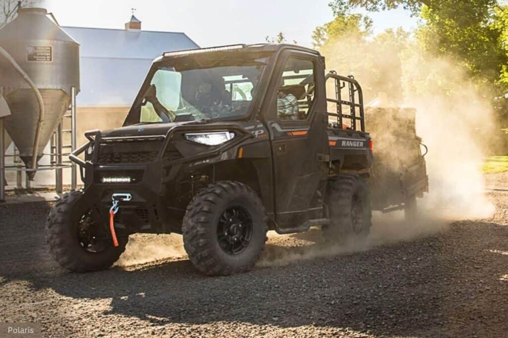 A Polaris Ranger UTV is driving on a gravel surface towing a trailer, generating a cloud of dust.
