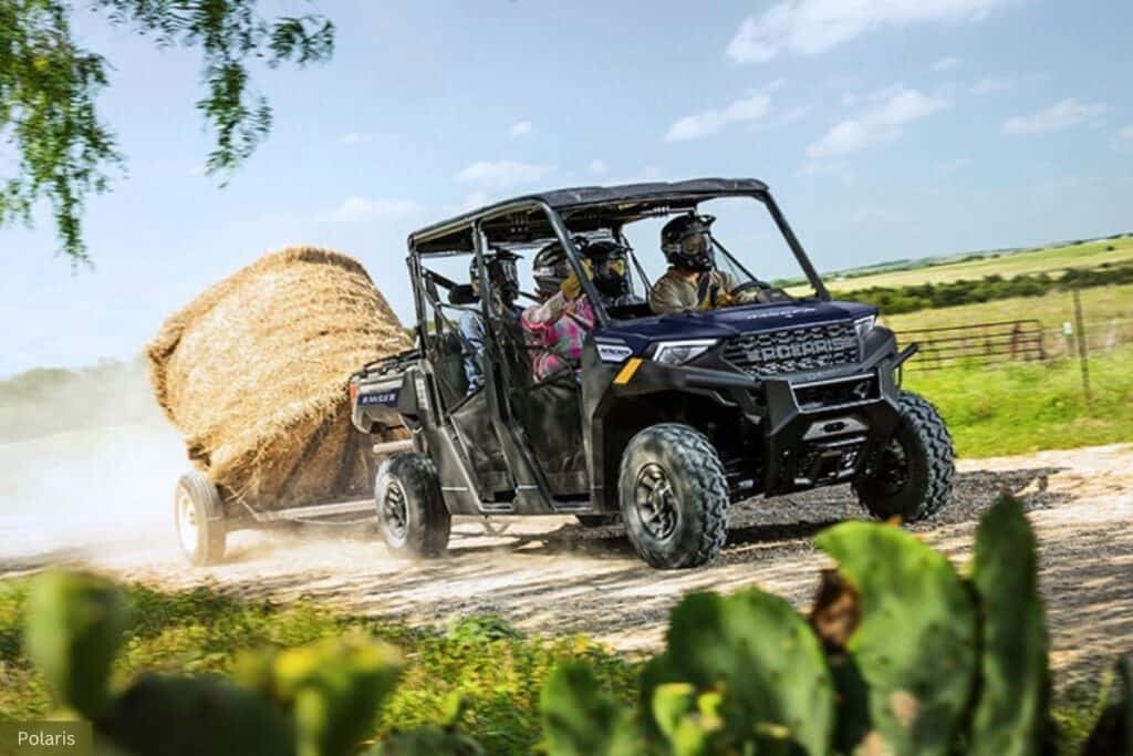 Side-by-side in motion, kicking up dust as it travels along a dirt path. The UTV, which is a Polaris Ranger, is towing a large round hay bale on a trailer.