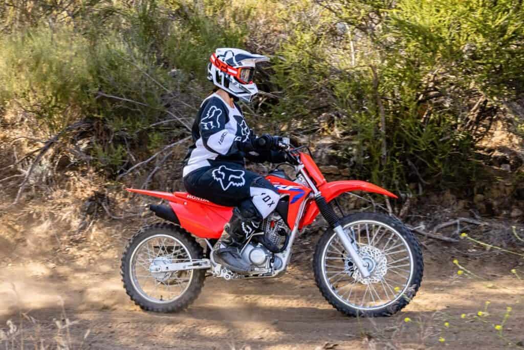 A rider on a Honda CRF125F dirt bike navigating a trail through sparse woodland.
