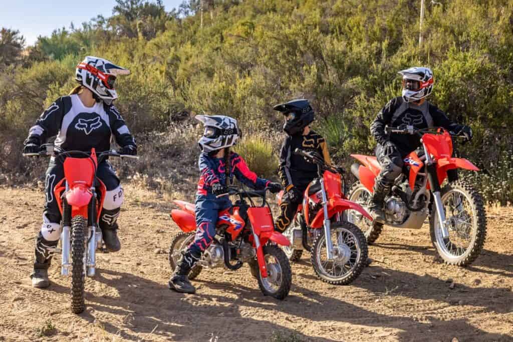A group of riders on Honda dirt bikes of varying sizes and engine displacements, including small models for children and larger ones for adults, ready for a ride on a dirt trail.