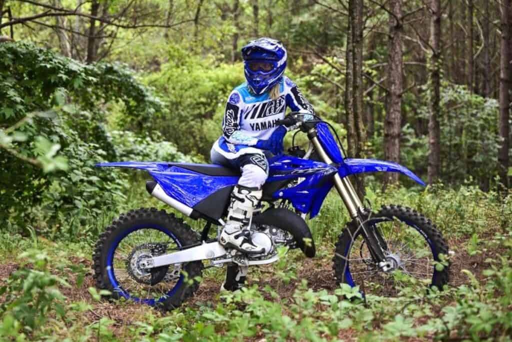 A rider dressed in full racing gear is on a blue Yamaha dirt bike, stopped in a lush green forest.