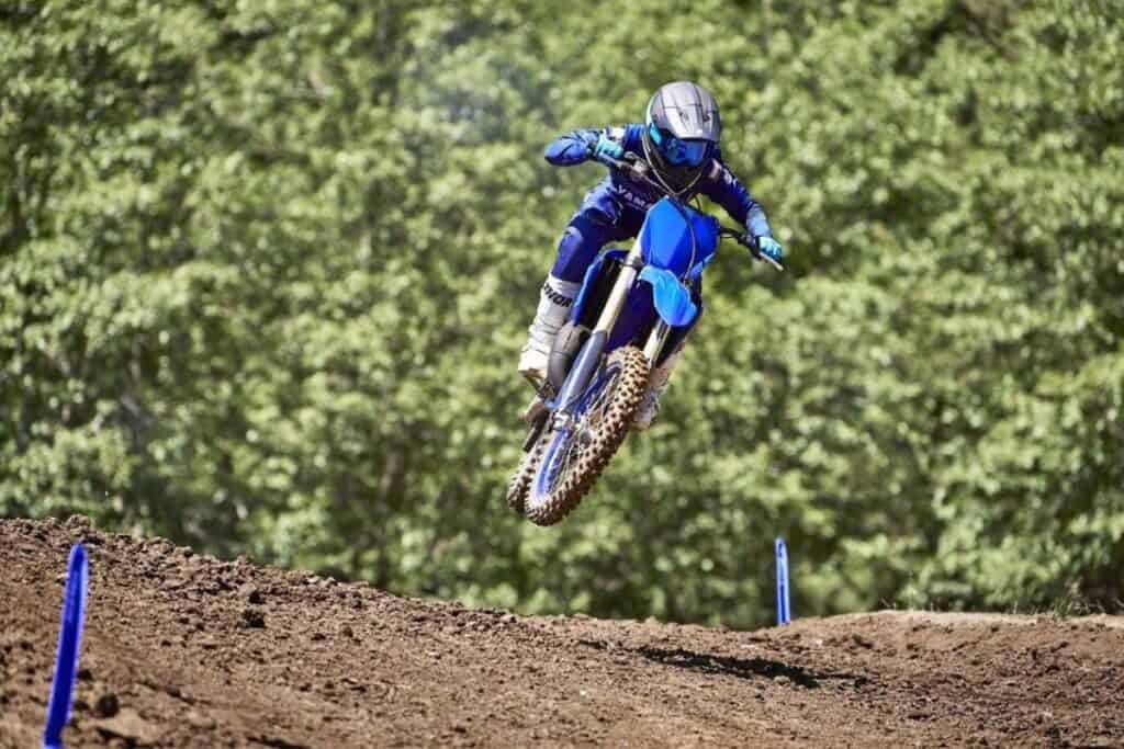 Motocross rider jumping a blue Yamaha YZ125 dirt bike on a dirt track surrounded by trees.