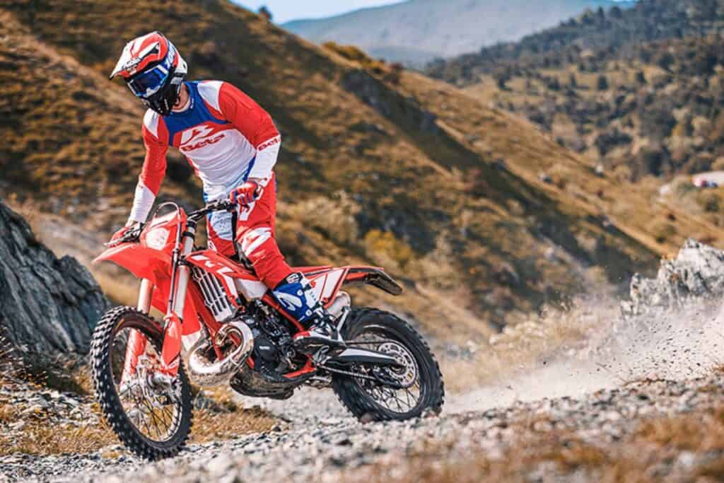 Amidst rugged terrain, a rider dressed in red and blue rides a Beta 300 RR dirt bike along a rocky mountain trail.