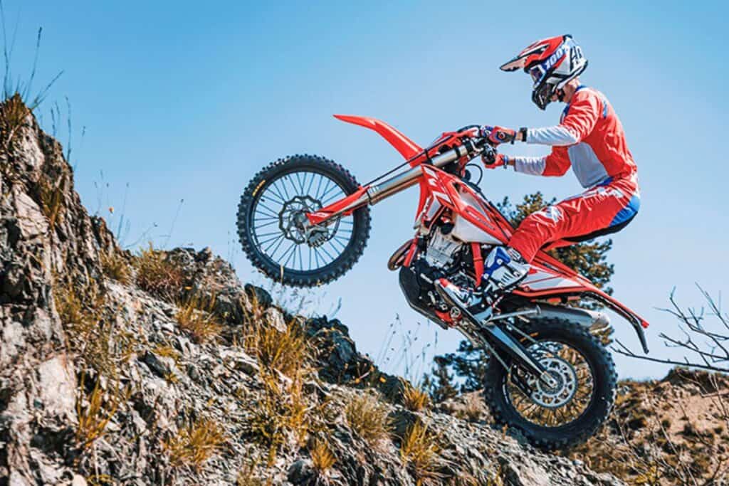 A rider in red and blue gear performing a wheelie on a Beta 300 RR dirt bike on a rocky hillside.