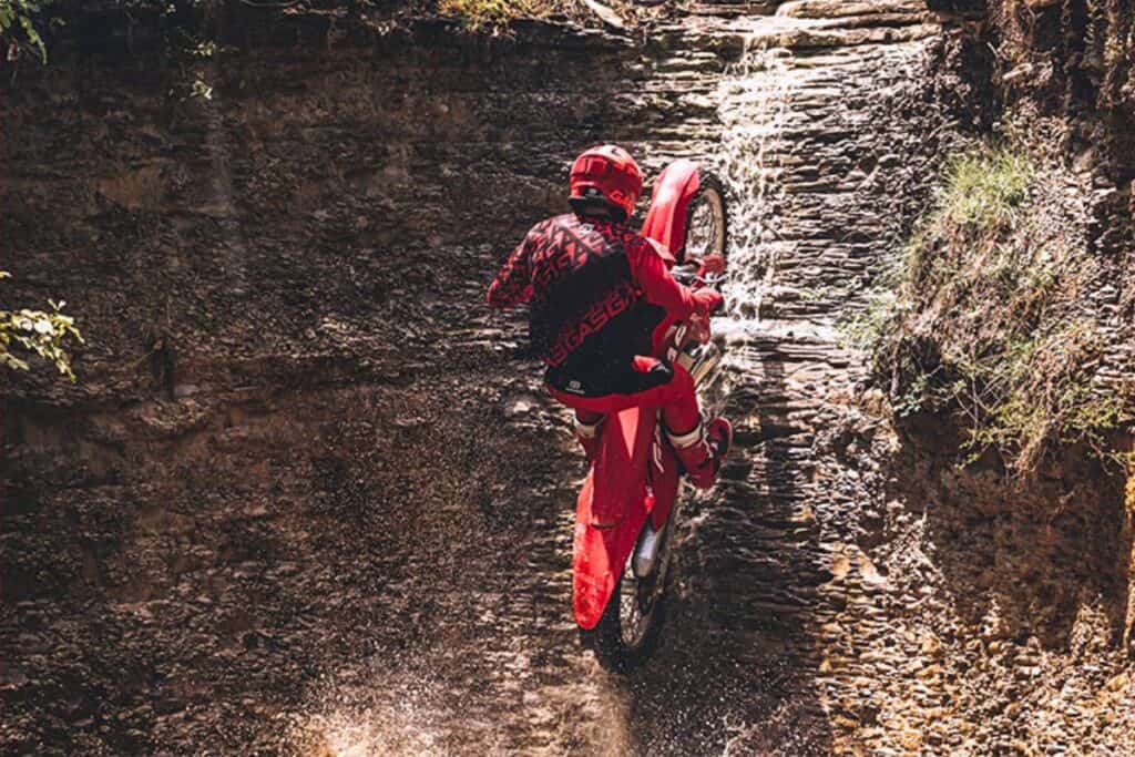 Climbing a small waterfall, a rider in red maneuvers a GasGas EC 300, creating a splash underneath.
