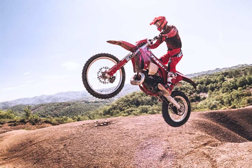A rider dressed in red launches a GasGas EC 300 dirt bike into the air on a dirt trail with a mountainous landscape in the background.