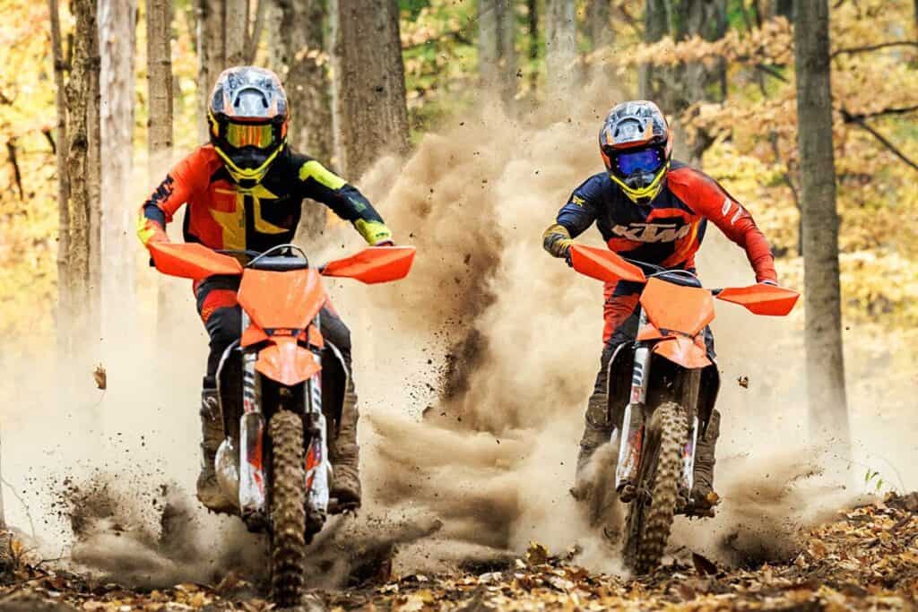 Competitors fiercely race their KTM 300 XC dirt bikes, churning up a cloud of dust on a forest trail carpeted with fallen leaves.