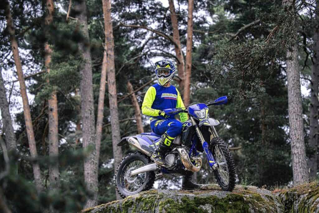 Poised on a rocky ledge, a rider in blue and neon gear surveys the trail on a Sherco 300 SE Factory dirt bike, surrounded by forest greenery.