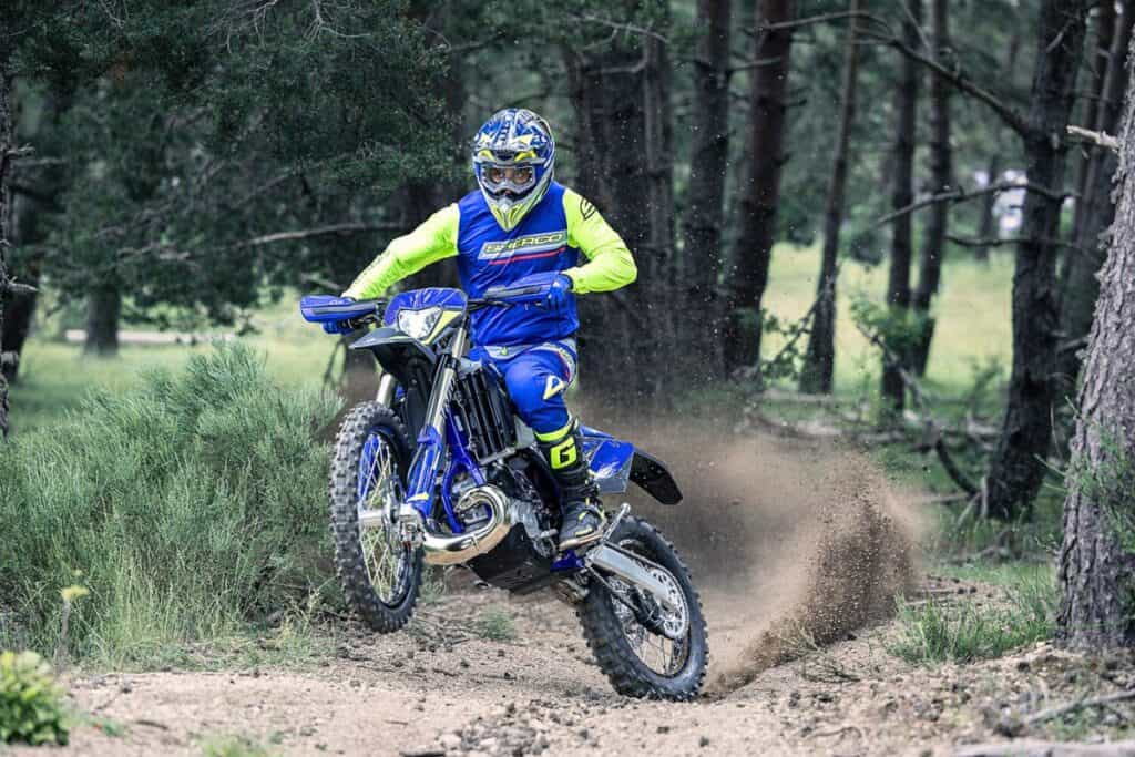 A rider in blue and fluorescent gear is riding a Sherco 300 SE Factory dirt bike through a forest trail, kicking up a trail of dust.