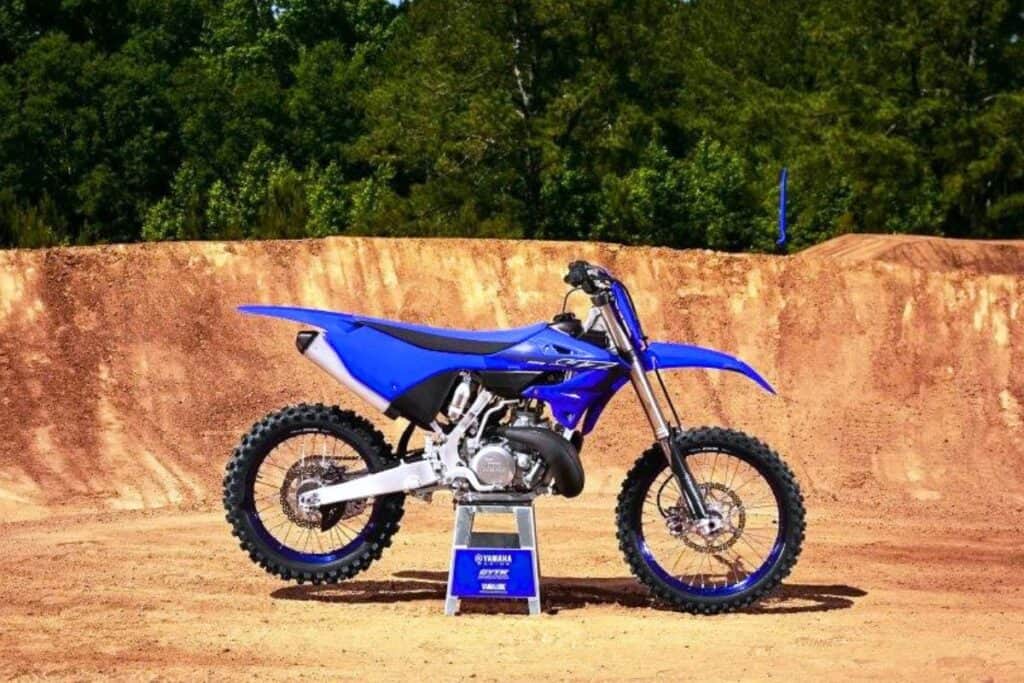 A Yamaha dirt bike is displayed on a stand in the center of a dirt track, with a backdrop of lush trees.