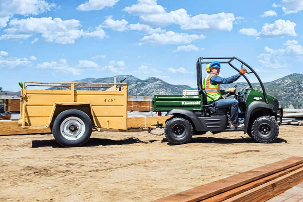 A construction worker driving a Kawasaki Mule SX UTV towing a trailer on a building site.