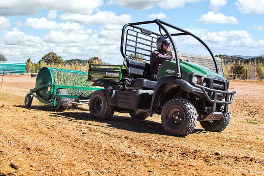 A rider driving a Kawasaki Mule SX UTV towing a spreader on dry land.