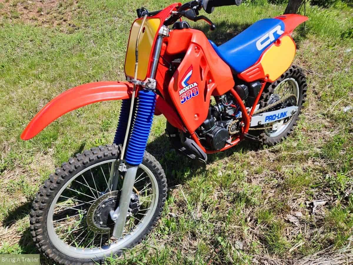 A Honda CR500 dirt bike with a yellow blank number plate, red body work, and a blue seat, standing in a grassy area.