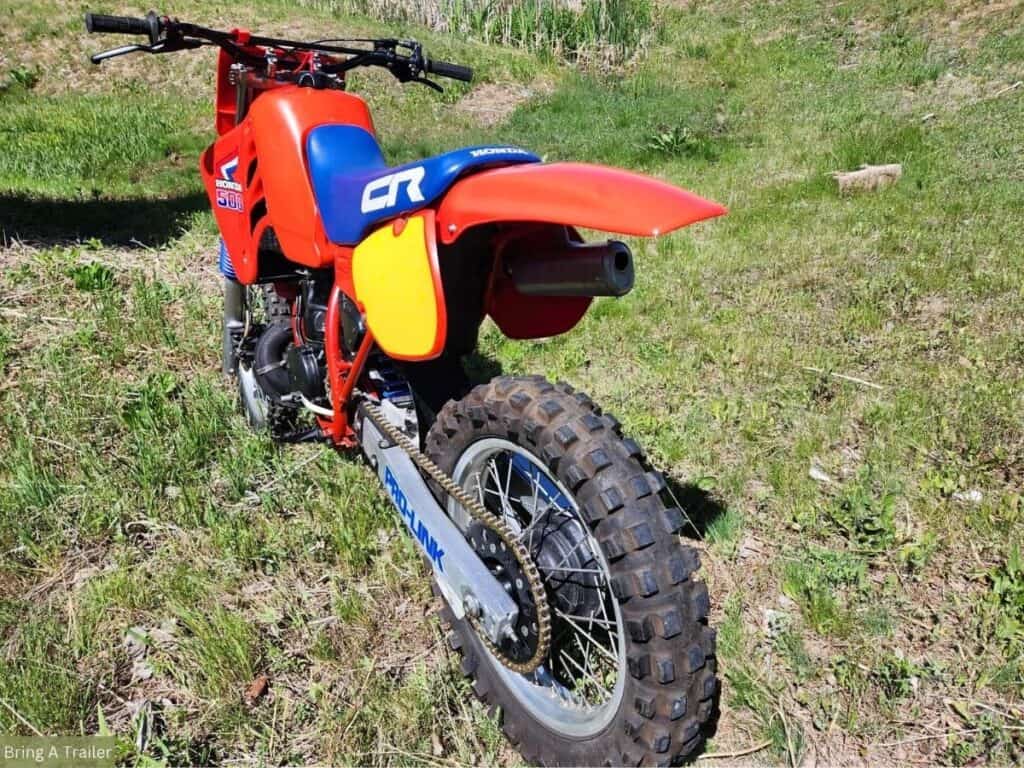 The rear view of a Honda CR500 dirt bike with a red and blue color scheme, parked in a grassy field.