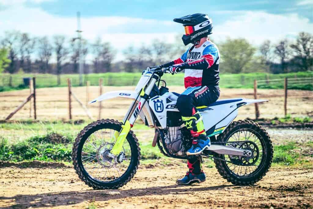 A dirt bike rider in full gear sitting on a white and blue dirt bike in a rural field with a wooden fence and trees in the background.