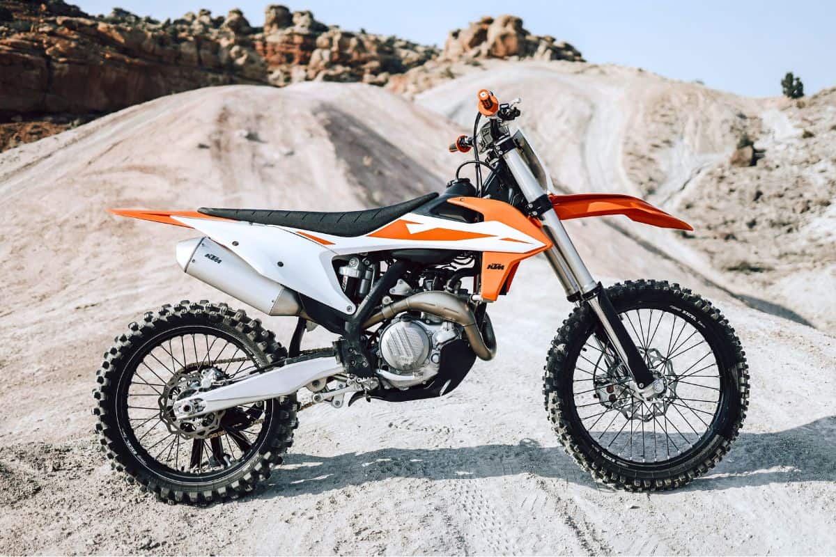 A KTM dirt bike with white and orange coloring, positioned on a desert trail with rocky terrain in the background.