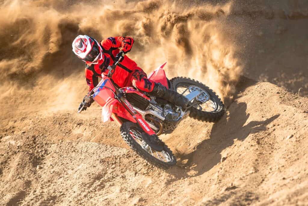 A rider in red motocross gear aggressively corners a  Honda CRF450R on a dusty track, kicking up a large cloud of dirt.