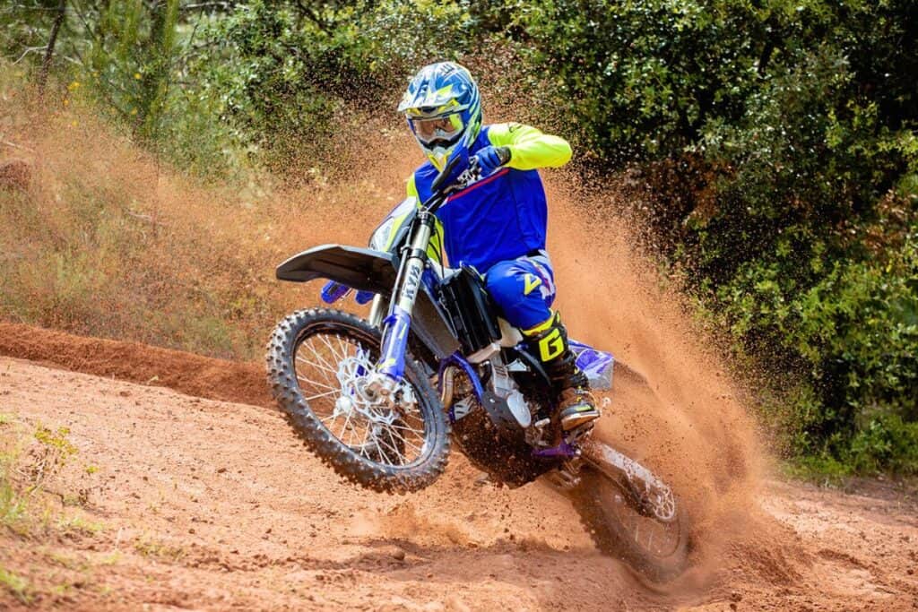 A rider in blue and yellow gear is aggressively riding a Sherco 450 SEF dirt bike, carving through a dirt track with a spray of dust trailing behind.